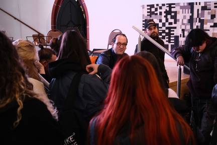 Treppe vor Bühne. eine Schulklasse, von hinten fotografiert, steht davor und hat fragen. Lukas sitzt und schaut richting kamera