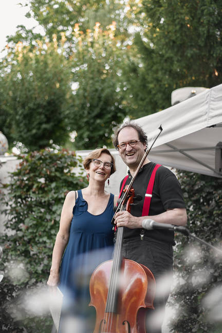 Sylvia Bachmann - Sprecherin (links) mit Lukas Dreyer - Cellist (rechts), beide lachend, in der Mitte das Cello im Vordergrund verschwommene weiße Blüten, Kulisse: ein Pavillon rechts hinten mit Hecken und Bäumen